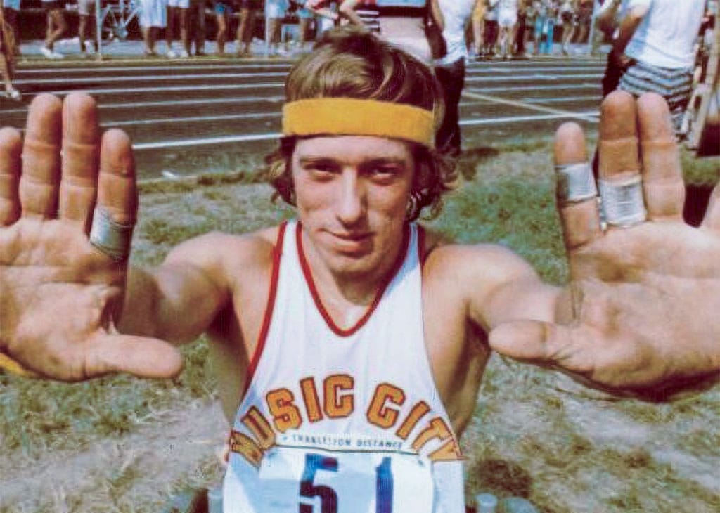 Bob Hall at the Boston Marathon in the mid-1970s, wearing a racing singlet and headband, holding his hands up toward the camera on the infield.