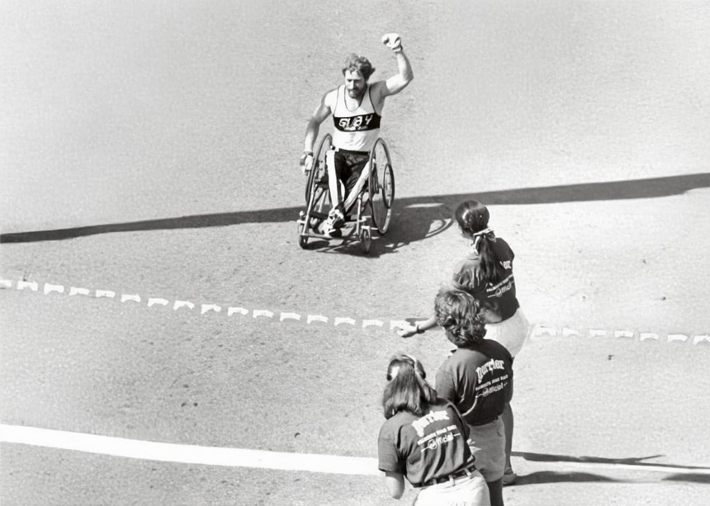 Bob Hall crosses the Marathon finish line in a racing wheelchair, raising his arm as spectators look on.