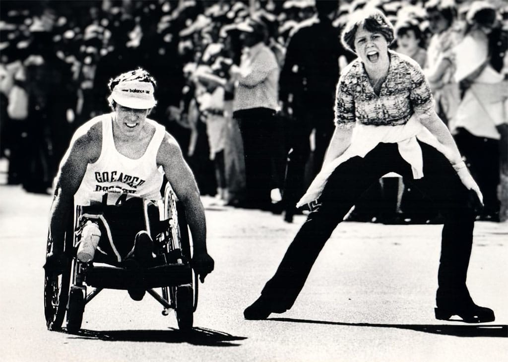 Bob Hall competing in a wheelchair on the Boston Marathon course, alongside his sister who is running and cheering beside him as spectators line the street.