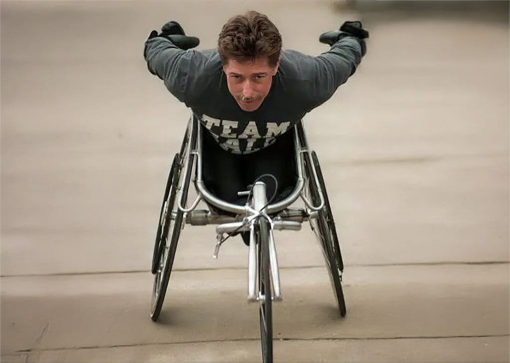 Bob Hall leaning forward in a racing wheelchair with his arms extended behind him, wearing a Team Hall shirt on a paved surface.