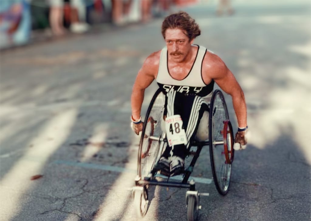 Bob Hall competing in a wheelchair during the Boston Marathon, pushing uphill on the course while wearing a numbered racing singlet.